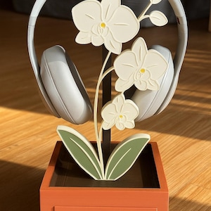May include: A decorative headphone stand featuring a white orchid design. The stand has a brown metal frame, a square orange base with a drawer, and holds a pair of gray headphones. The orchid has white flowers with yellow centers and green leaves.
