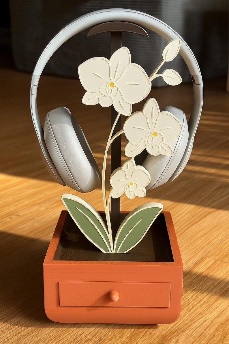 May include: A decorative headphone stand featuring a white orchid design. The stand has a brown metal frame, a square orange base with a drawer, and holds a pair of gray headphones. The orchid has white flowers with yellow centers and green leaves.
