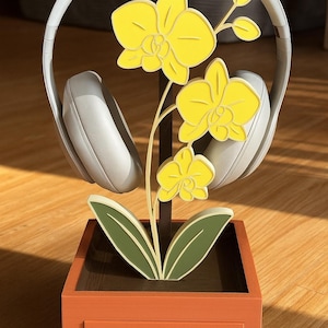 May include: A decorative headphone stand featuring a yellow orchid design. The stand is orange with a small drawer and holds a pair of white headphones. The orchid has yellow flowers and green leaves, adding a touch of nature to the design.