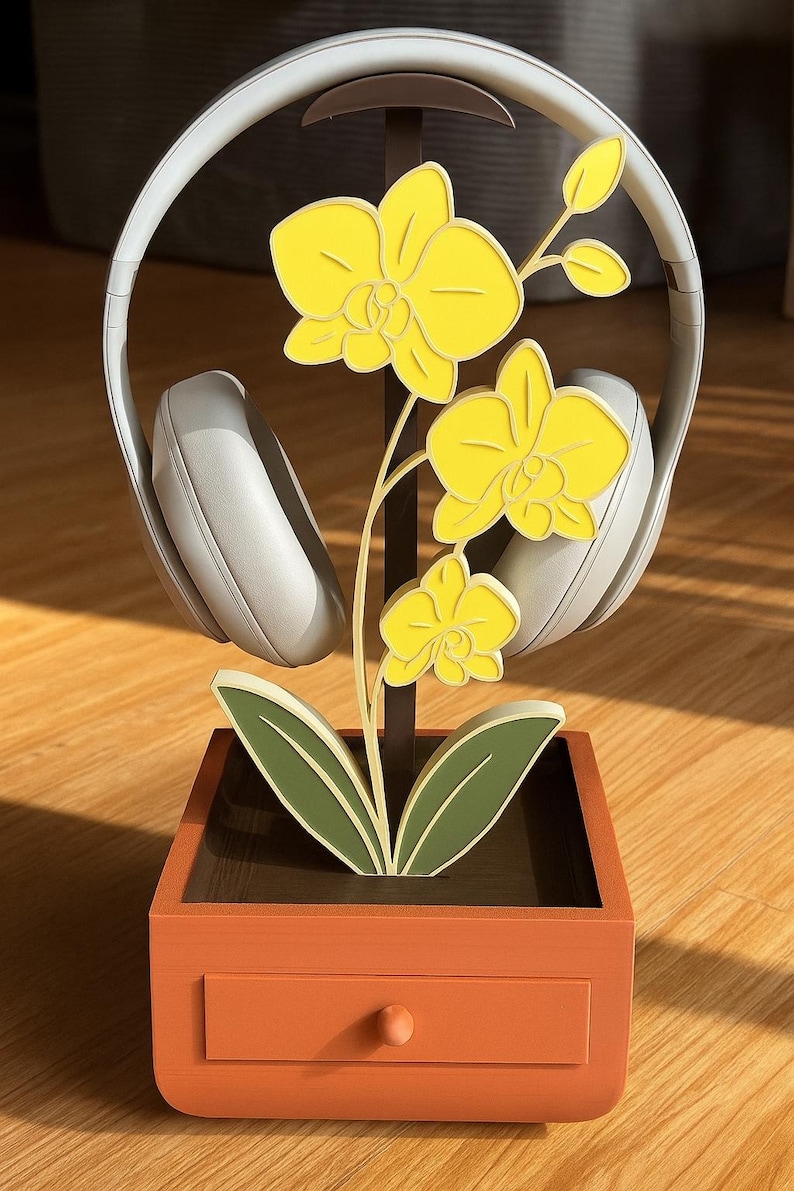 May include: A decorative headphone stand featuring a yellow orchid design. The stand is orange with a small drawer and holds a pair of white headphones. The orchid has yellow flowers and green leaves, adding a touch of nature to the design.