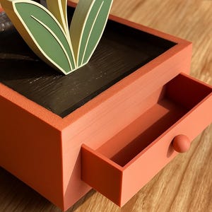 May include: A decorative orange-red box with a drawer, featuring a stylized flower with green leaves and pink petals. The drawer has a small, round knob. The box sits on a wooden surface.