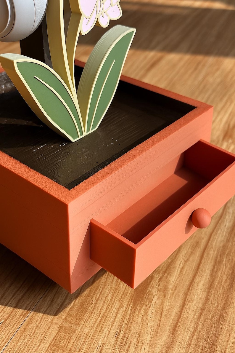 May include: A decorative orange-red box with a drawer, featuring a stylized flower with green leaves and pink petals. The drawer has a small, round knob. The box sits on a wooden surface.