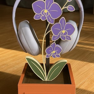 May include: A decorative headphone stand featuring a purple orchid design. The stand is made of wood and has a drawer. Gray headphones rest on the stand. The orchid has purple flowers with yellow centers and green leaves.