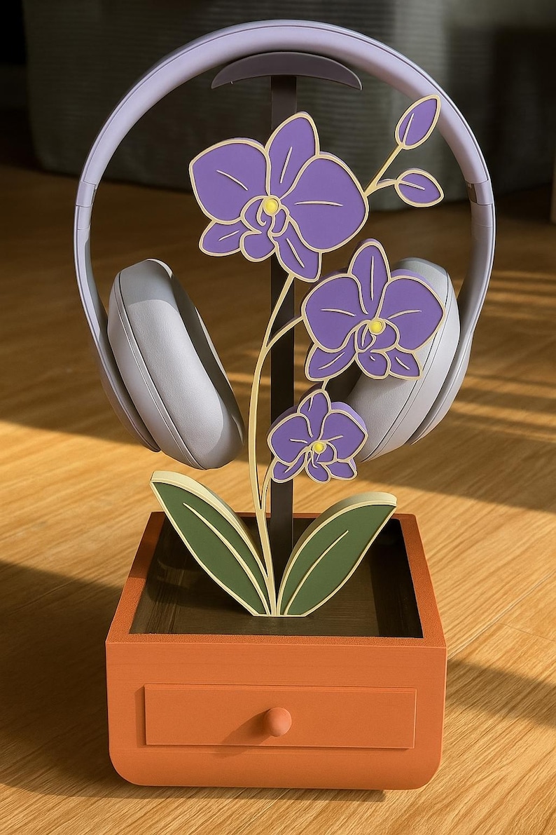May include: A decorative headphone stand featuring a purple orchid design. The stand is made of wood and has a drawer. Gray headphones rest on the stand. The orchid has purple flowers with yellow centers and green leaves.