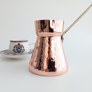 May include: A polished copper coffee pot with a hammered texture and a long, slender metal handle, positioned next to a white ceramic cup and saucer featuring a detailed blue, red, and green floral pattern with a decorative rim design.