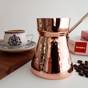 May include: A hammered copper Turkish coffee pot with a long handle. The pot is sitting on a white surface with a cup of coffee and a box of Turkish Delight in the background.