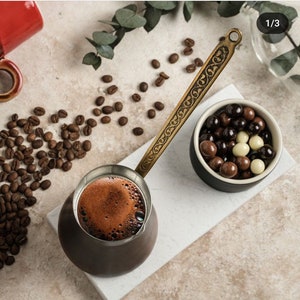 May include: A traditional Turkish coffee pot with a brass handle and intricate design. The pot is filled with dark coffee and sits on a white marble surface. A small bowl filled with chocolate sweets is also visible.
