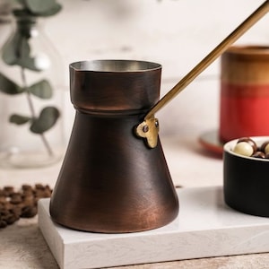 May include: A copper and brass Turkish coffee pot with a long handle. The pot is sitting on a white surface.