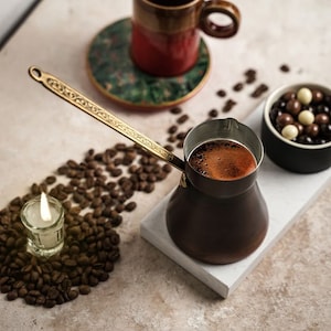 May include: A copper coffee pot with a decorative gold handle, filled with coffee, sits on a white tray. Coffee beans surround a small candle and a cup of coffee. A small bowl of chocolate sweets is also present.