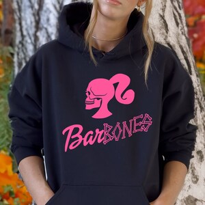 Op de afbeelding: Een zwarte hoodie met een roze schedel en de tekst "BarBones" erop gedrukt. De hoodie wordt gedragen door iemand die op een pompoen zit in een herfstachtige omgeving.