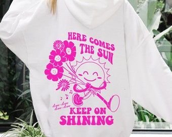 May include: White hooded sweatshirt with a pink graphic of a smiling sun holding a bunch of flowers and the text "Here comes the sun, doo-doo-doo-doo keep on shining."
