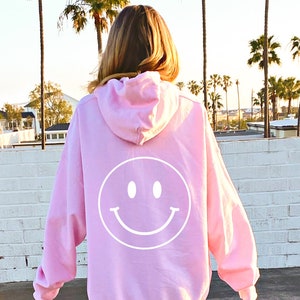 May include: A pink hoodie with a white smiley face on the back. The hoodie is oversized and has a drawstring hood.