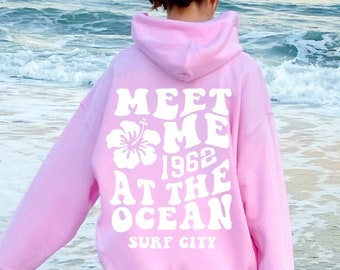May include: A pink hoodie with a white graphic that says "Meet Me 1962 At The Ocean Surf City" with a white hibiscus flower graphic. The person wearing the hoodie is walking in the ocean.