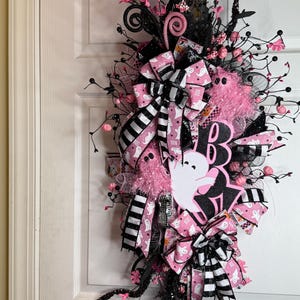 May include: A Halloween wreath in black and pink. It features a 'Boo' sign, ghost cutouts, and bows with a ghost pattern. The design includes pink and black accents, faux flowers, and decorative branches, creating a festive look.