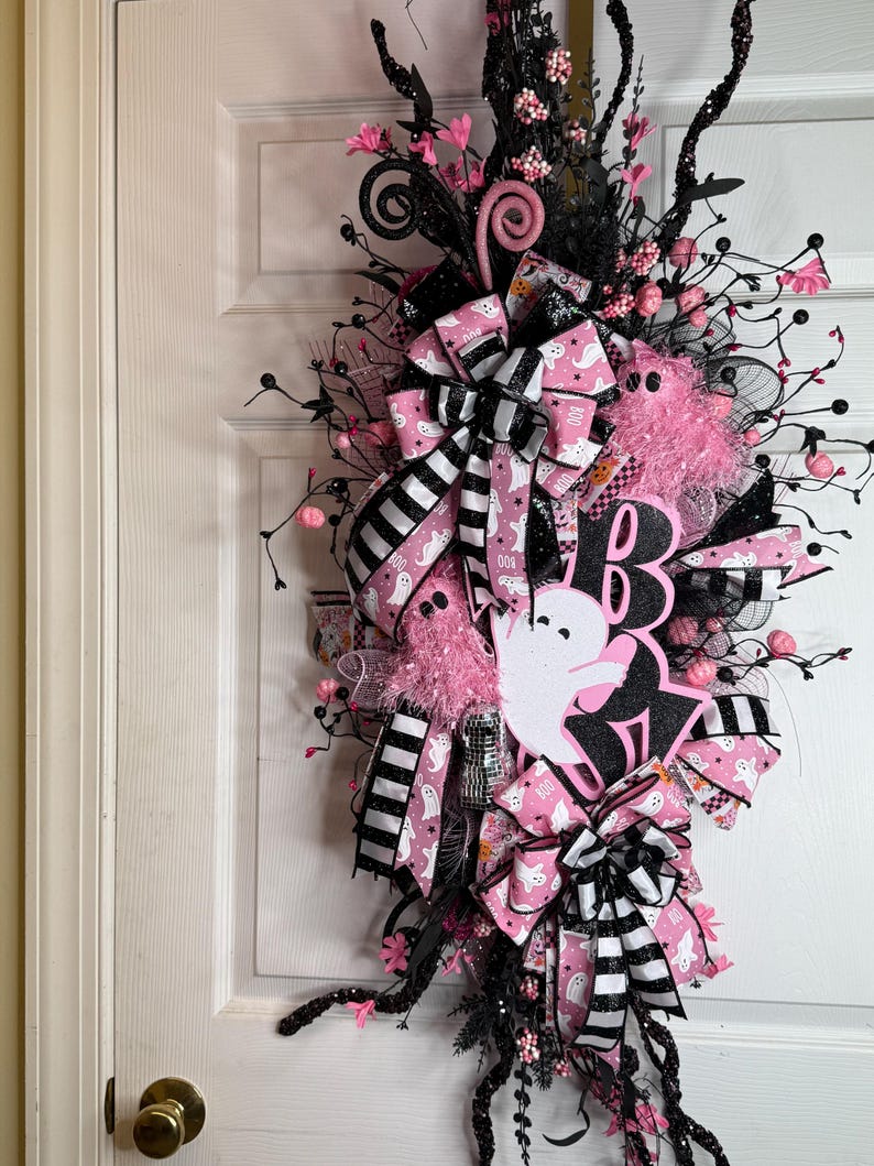 May include: A Halloween wreath in black and pink. It features a 'Boo' sign, ghost cutouts, and bows with a ghost pattern. The design includes pink and black accents, faux flowers, and decorative branches, creating a festive look.