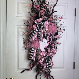May include: A Halloween wreath with a vertical design, featuring black and pink ribbons, bows, and decorative elements. The wreath includes a white ghost cutout and the word "BOO" in black and pink. The wreath is adorned with pink flowers and black branches.