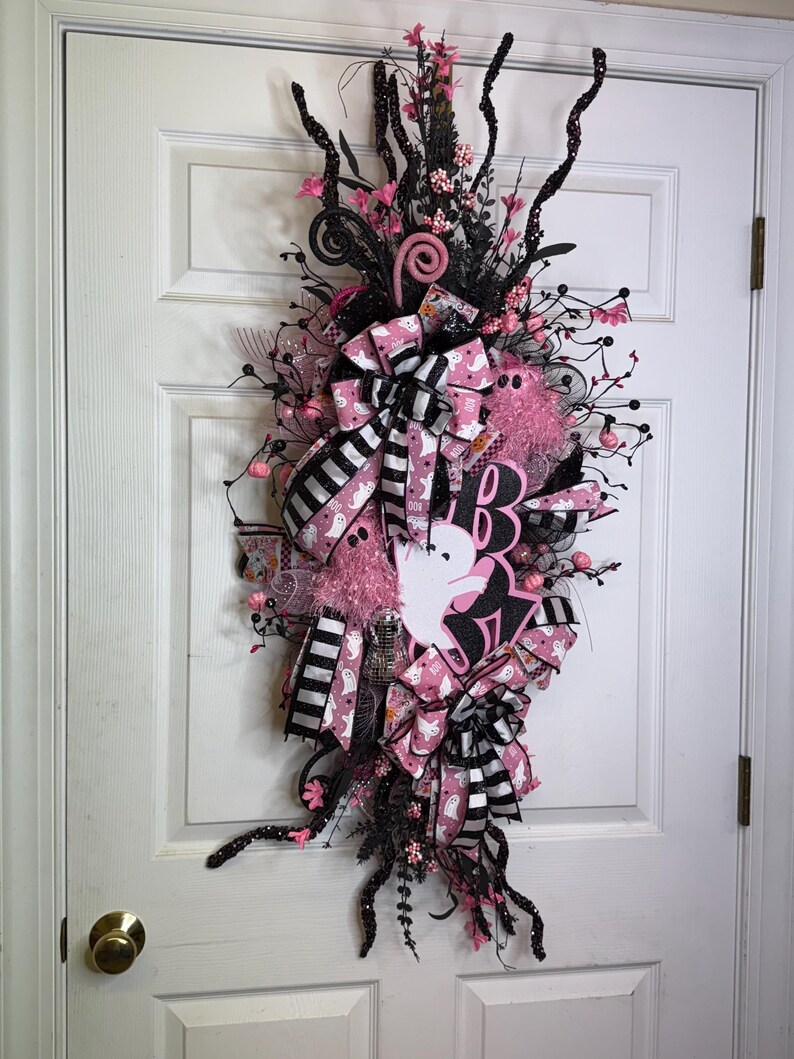 May include: A Halloween wreath with a vertical design, featuring black and pink ribbons, bows, and decorative elements. The wreath includes a white ghost cutout and the word "BOO" in black and pink. The wreath is adorned with pink flowers and black branches.