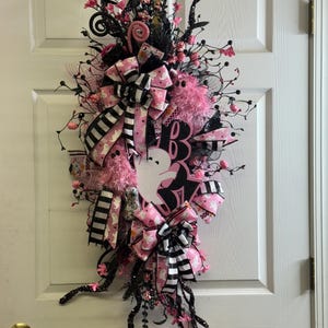 May include: A Halloween wreath with a white ghost silhouette, pink and black ribbons, and decorative floral accents. The wreath features the word "Boo" and is adorned with black branches and pink embellishments.