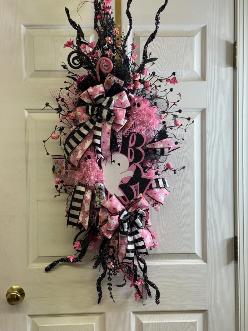 May include: A Halloween wreath with a white ghost silhouette, pink and black ribbons, and decorative floral accents. The wreath features the word "Boo" and is adorned with black branches and pink embellishments.