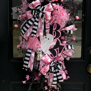 May include: A Halloween wreath with a black base, decorated with pink and black ribbons, bows, and floral details. Includes a white ghost cutout and the word "BOO" in pink. Festive and decorative.