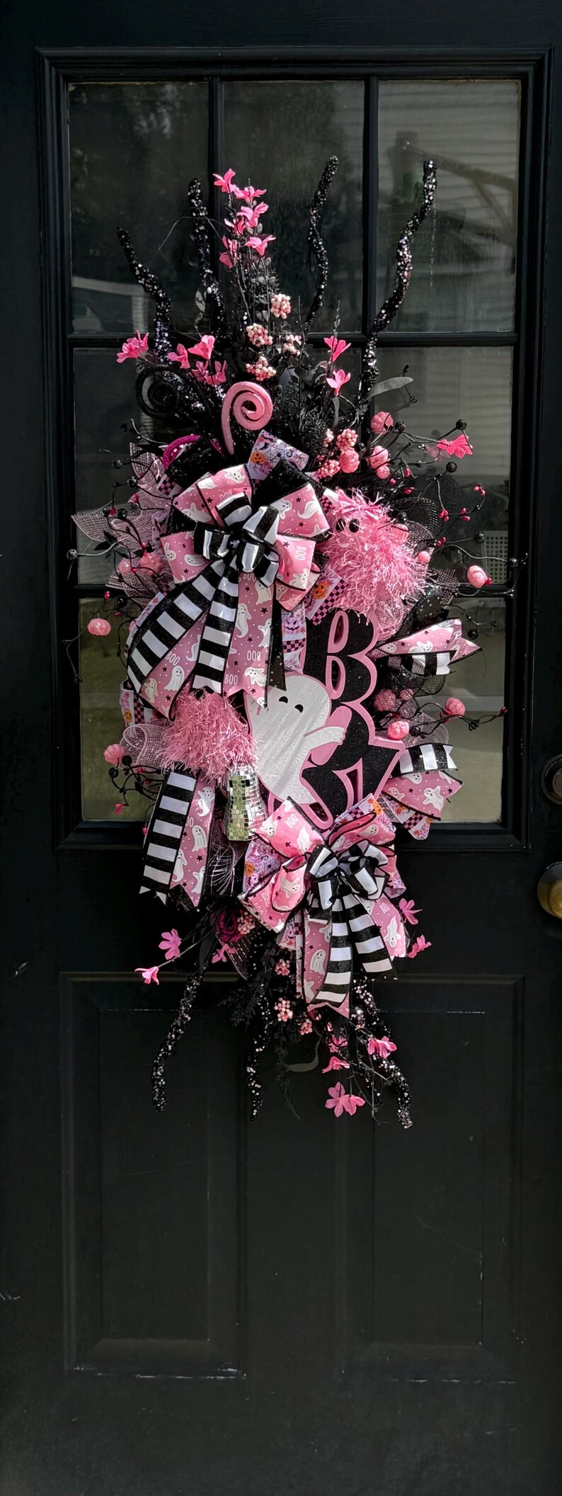 May include: A Halloween wreath with a black base, decorated with pink and black ribbons, bows, and floral details. Includes a white ghost cutout and the word "BOO" in pink. Festive and decorative.