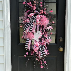 May include: A Halloween wreath with pink and black decorations, including a ghost and the letter "B". The wreath has pink bows, decorative flowers, and striped ribbons. It is hanging on a black door.