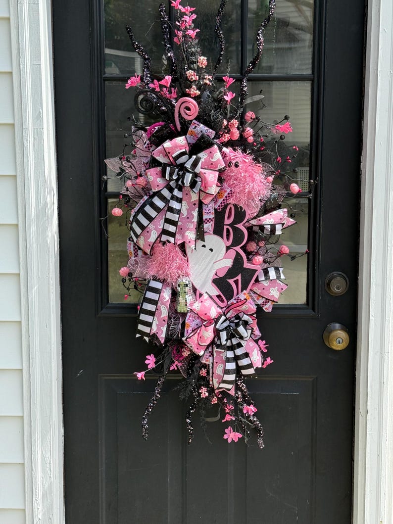 May include: A Halloween wreath with pink and black decorations, including a ghost and the letter "B". The wreath has pink bows, decorative flowers, and striped ribbons. It is hanging on a black door.