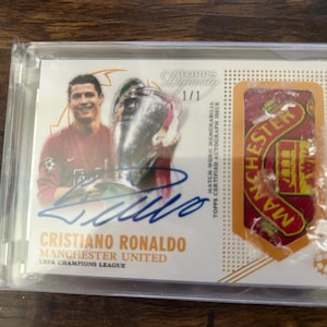 Puede incluir: Tarjeta coleccionable con Cristiano Ronaldo con la camiseta del Manchester United, sosteniendo un trofeo. La tarjeta incluye un autógrafo y el logotipo del Manchester United. La tarjeta está en un soporte protector.