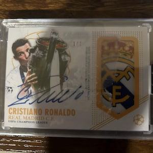 May include: A collectible trading card featuring Cristiano Ronaldo, holding a trophy, with his signature in blue ink. The card also displays the Real Madrid C.F. logo and the text "UEFA Champions League."