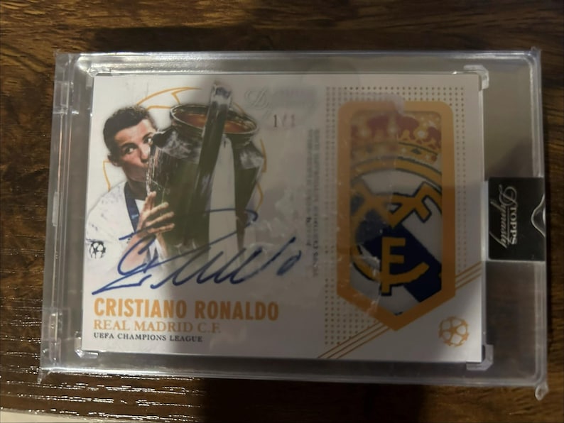 May include: A collectible trading card featuring Cristiano Ronaldo, holding a trophy, with his signature in blue ink. The card also displays the Real Madrid C.F. logo and the text "UEFA Champions League."