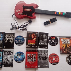 May include: A red and black electric guitar video game controller with a strap, along with a collection of PlayStation 2 video game discs and cases, including Rock Band, Rock Band 2, AC/DC Live Track Pack, Guitar Hero III Legends of Rock, and Guitar Hero III Legends of Rock.