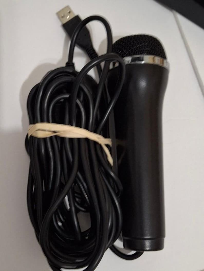 May include: A black USB microphone with a silver rim around the mesh head. The microphone has a black cable that is coiled and secured with a rubber band. The USB connector is visible.
