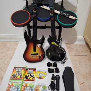 May include: A complete Guitar Hero and Band Hero game set. Includes two guitar controllers, a drum set, game discs, and accessories. The guitars have black bodies with colorful fret buttons. The drum set has black pads with colored rims.