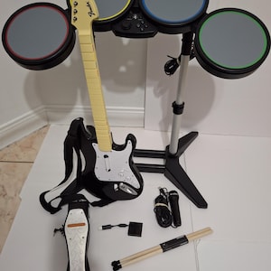 May include: A black and white Fender Stratocaster guitar with a cream-colored neck rests against a drum set with gray pads and colored rims. Accessories include a foot pedal, drumsticks, a microphone, and connecting cables.