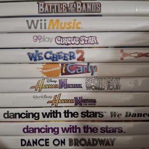 May include: A stack of Wii game cases featuring titles like "Battle of the Bands," "Wii Music," and "We Cheer 2." Other titles include "iCarly," "Hannah Montana," and "Dancing with the Stars." The cases are white with colorful text and graphics.
