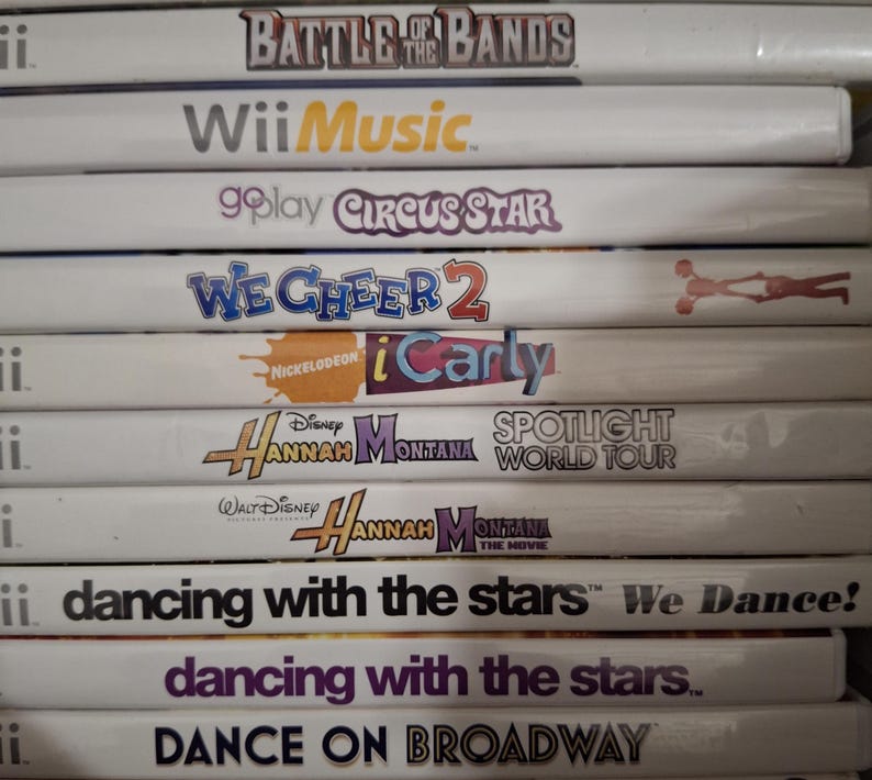 May include: A stack of Wii game cases featuring titles like "Battle of the Bands," "Wii Music," and "We Cheer 2." Other titles include "iCarly," "Hannah Montana," and "Dancing with the Stars." The cases are white with colorful text and graphics.