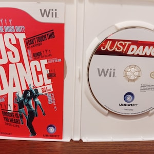 May include: Open Wii game case featuring the game "Just Dance." The red cover displays song titles and dancing figures. The disc inside has the game title and Wii logo. A fun video game for all ages.