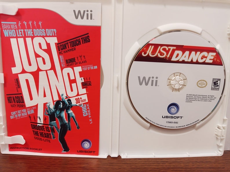 May include: Open Wii game case featuring the game "Just Dance." The red cover displays song titles and dancing figures. The disc inside has the game title and Wii logo. A fun video game for all ages.