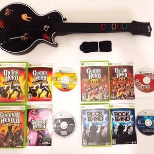 May include: A black Guitar Hero guitar controller with colorful fret buttons and a black strap. The image also shows several Xbox 360 game cases and discs, including Guitar Hero and Rock Band titles.