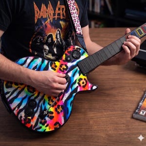 May include: A colorful guitar controller with a rainbow zebra and leopard print design. The guitar has a black neck and buttons. The strap is pink with a leopard print. The person is wearing a black t-shirt with a band graphic.