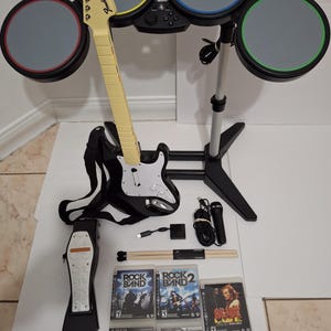 May include: A Rock Band video game set, including a drum set, guitar, microphone, and foot pedal. The set includes several game discs, drumsticks, and connecting cables. The guitar is cream-colored with a black body.
