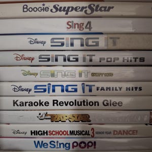 May include: A stack of video game cases featuring titles like "Boogie SuperStar", "Sing 4", and several "Sing It" editions from Disney. Other titles include "Karaoke Revolution Glee" and "We Sing Pop!"