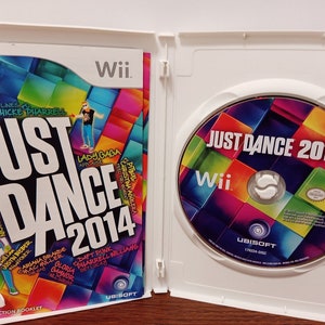 May include: An open white Wii game case featuring the game "Just Dance 2014." The game's cover art is colorful with the game title and artist names. The disc is visible on the right, also with the game title and Wii logo.