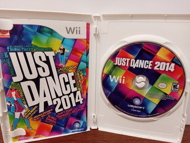 May include: An open white Wii game case featuring the game "Just Dance 2014." The game's cover art is colorful with the game title and artist names. The disc is visible on the right, also with the game title and Wii logo.