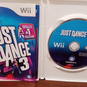 May include: A Wii game case for "Just Dance 3" featuring a blue and white design. The game disc is visible, along with the instruction booklet. The cover art includes the game title, artist names, and the Wii logo.