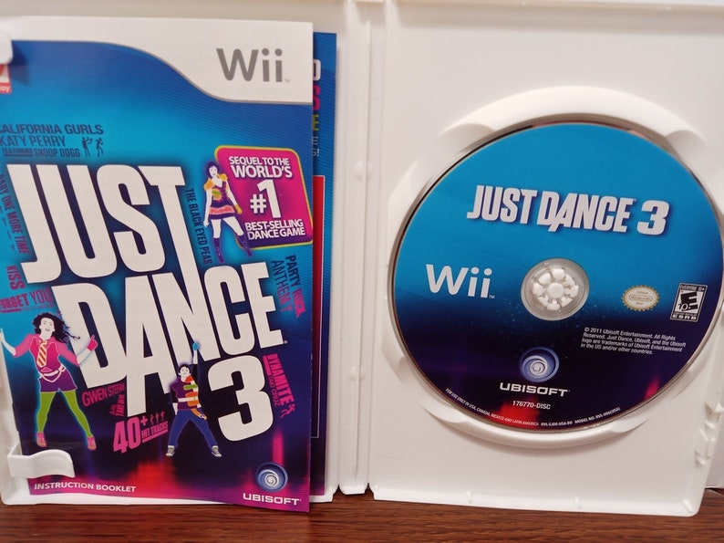May include: A Wii game case for "Just Dance 3" featuring a blue and white design. The game disc is visible, along with the instruction booklet. The cover art includes the game title, artist names, and the Wii logo.