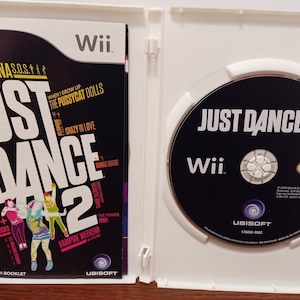 May include: A Wii game case for "Just Dance 2" featuring the game disc and instruction booklet. The booklet lists 40+ hit tracks, including songs by Rihanna, James Brown, and The Pussycat Dolls. The disc has the game title and Wii logo.