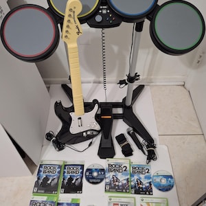 May include: A Rock Band video game set, including a black and white guitar, a drum set with gray, yellow, blue, and green pads, and several game discs. The set also includes a microphone, a foot pedal, and drumsticks.