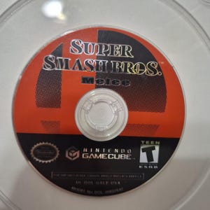 Może przedstawiać: Okrągła płyta z grą Nintendo GameCube do Super Smash Bros. Melee. Płyta ma czerwono-czarne tło z tytułem gry w białym i czarnym tekście, logo Nintendo GameCube oraz symbolem klasyfikacji wiekowej ESRB Teen.
