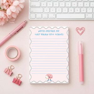 May include: A pink notepad with the text "NOTES BEFORE MY LAST BRAIN CELL LEAVES" in blue. The notepad has blue lines and a cartoon brain illustration. Pink pen, tape, paper clips, and a marker are also visible.